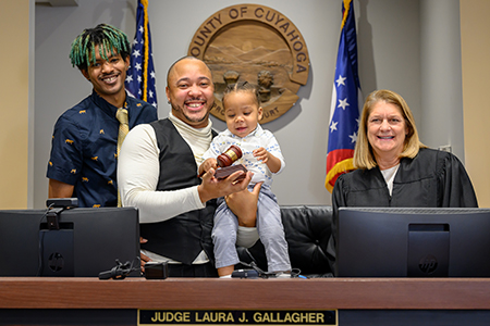 A family and a judge smile at a courtroom bench while a toddler holds a judge’s gavel during an adoption celebration.