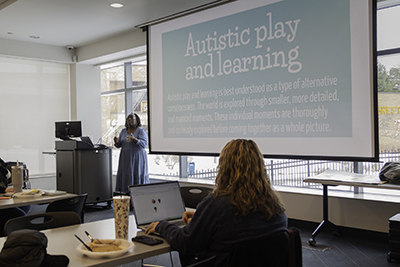 Milestones Training Universal Truths Presenter standing near a podium in a training room, speaking to attendees while a slide titled “Autistic Play and Learning” is projected on the screen.