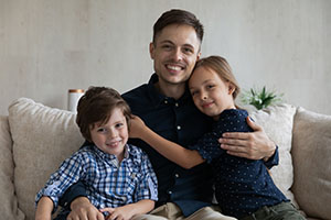 Father with son and daughter sitting together on sofa smile looking at camera.