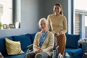 Cheerful multiethnic girl standing by her grandmather in wheelchair while both looking at camera with smiles against home environment