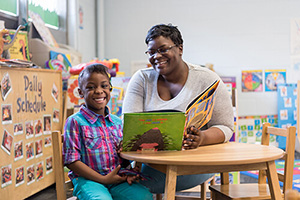 a woman and child sitting at a table and reading a book and smiling