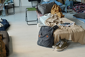 Gumshoes and backpack with refugee supplies standing by sleeper with knitted beige plaid, book of comics, headphones and smartphone